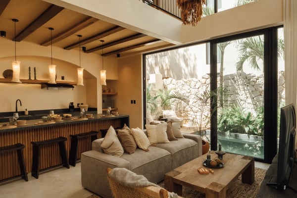 Covered wooden deck with woven chairs, potted palms, and sliding glass doors opening to modern kitchen with pendant lights